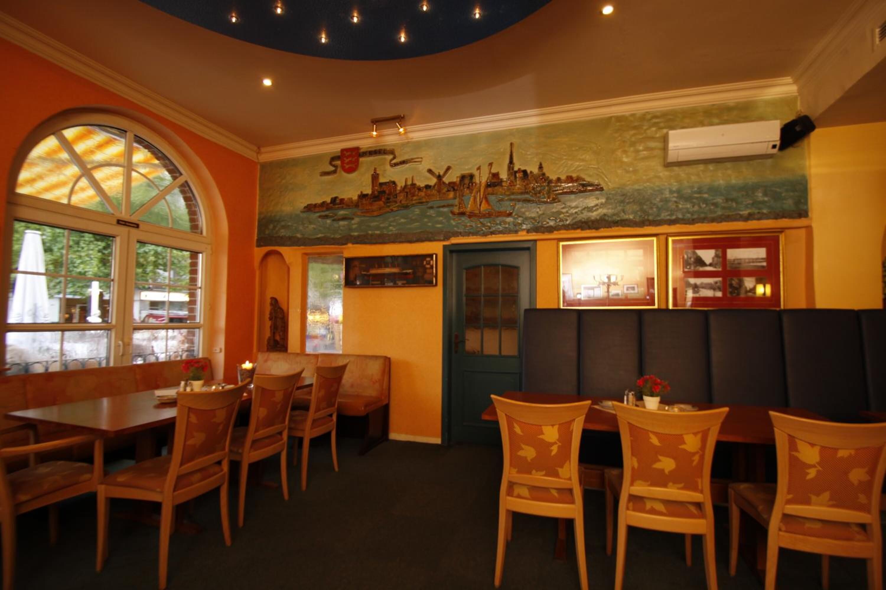 Interno di un ristorante con tavoli e sedie in legno, un affresco sulla parete e un'ampia finestra che si affaccia sull'esterno.