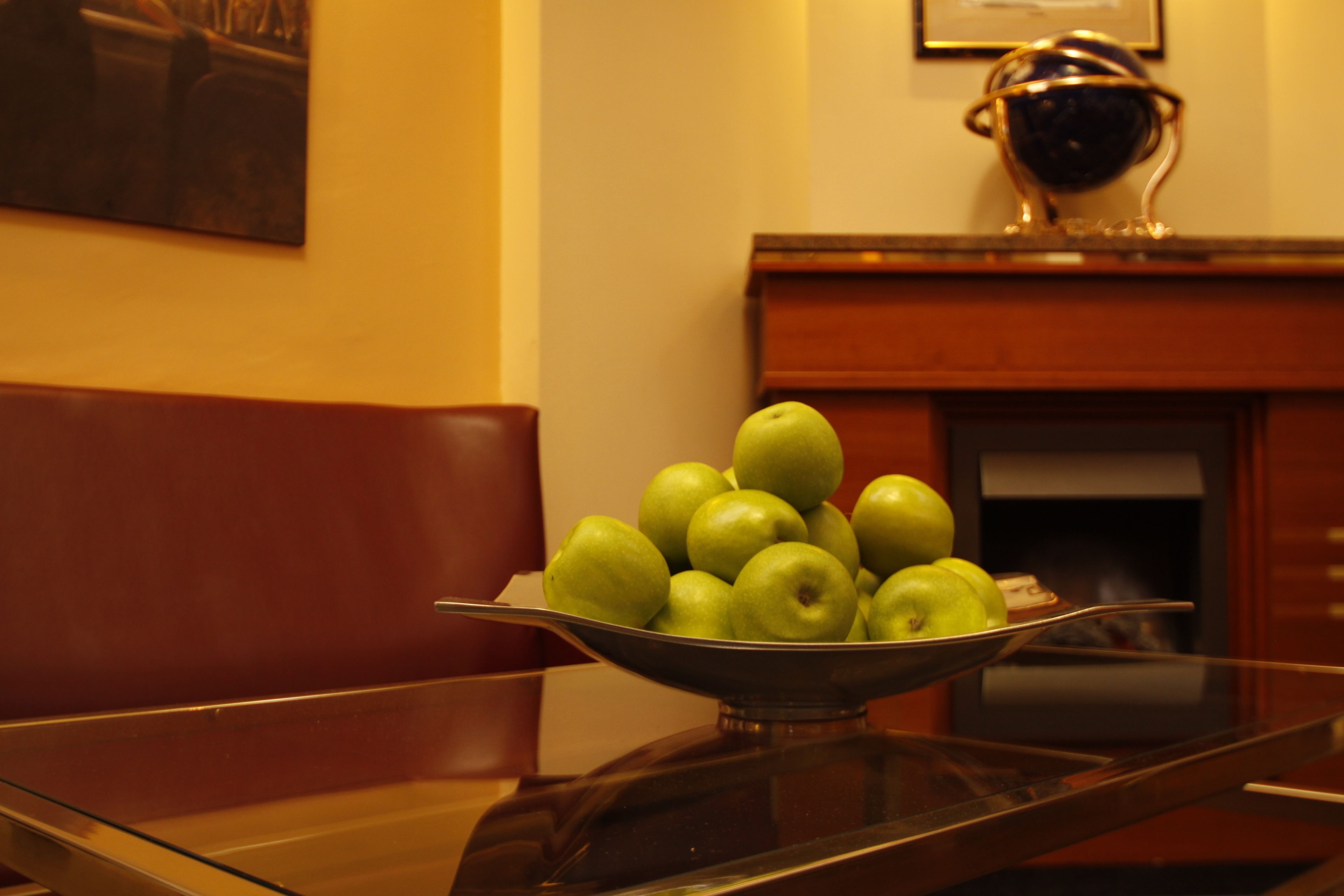 A bowl of green apples on a glass table with a brown leather chair and a fireplace in the background.