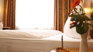 A hotel room with a neatly made bed, a vase with flowers, and a bowl of snacks on a wooden table. - Hotel Kaiserhof (Wesel)