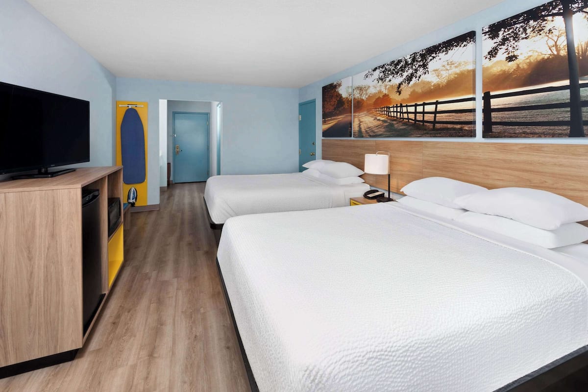 Una habitación de hotel con dos camas, un televisor, un armario de madera y un cuadro de un atardecer en la pared.
