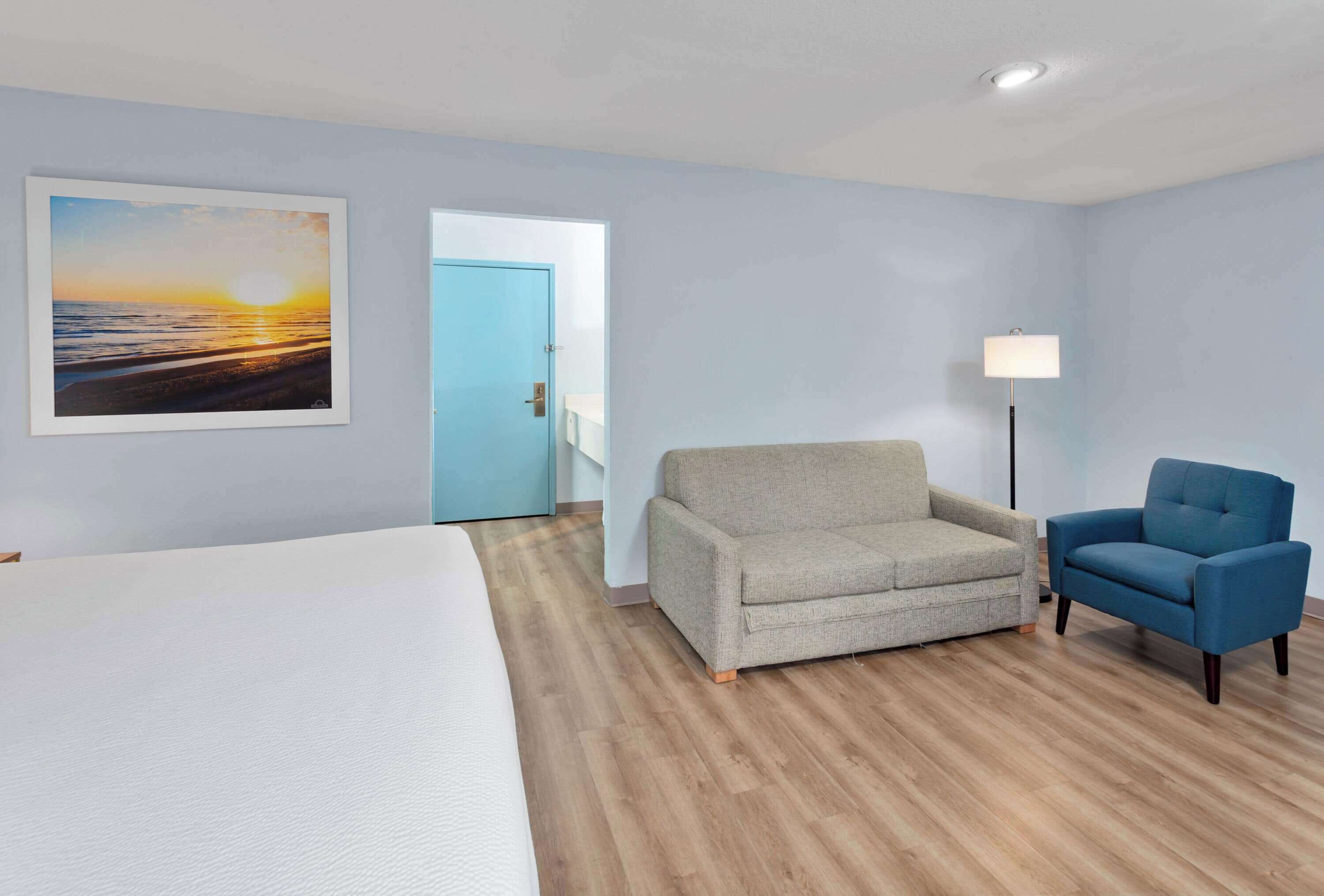 A hotel room with a bed, a grey sofa, a blue armchair, a painting of a sunset, and a door leading to another room.