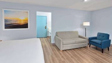 Une chambre d’hôtel avec un lit, un canapé gris, un fauteuil bleu, un tableau représentant un coucher de soleil et une porte donnant sur une autre pièce.