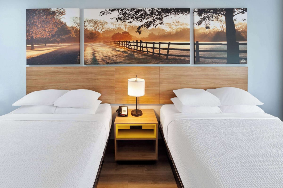 Habitación de hotel con dos camas, un cabecero de madera, una mesita de noche con lámpara y tres cuadros en la pared.