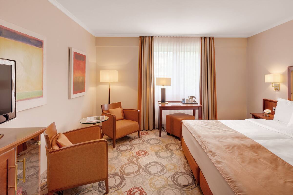 Une chambre d’hôtel comprenant un lit, deux fauteuils, un bureau, une télévision et une fenêtre avec des rideaux.