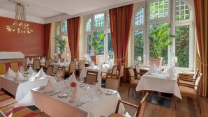 Restaurante com várias mesas preparadas para refeições, janelas amplas com cortinas e plantas dentro.