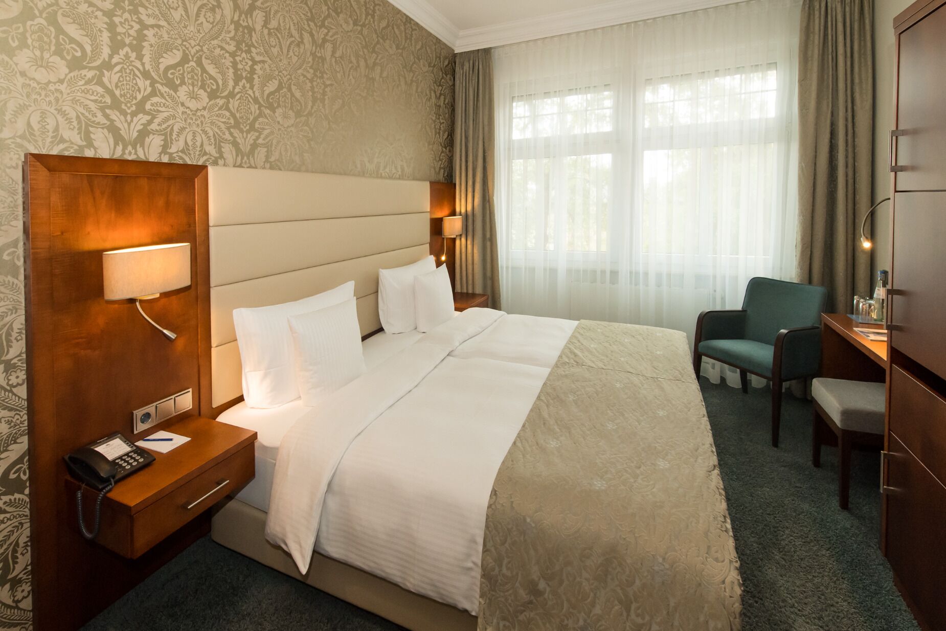 Habitación de hotel con una cama grande, lámparas de noche, un escritorio con una silla y vistas al exterior a través de cortinas traslúcidas.