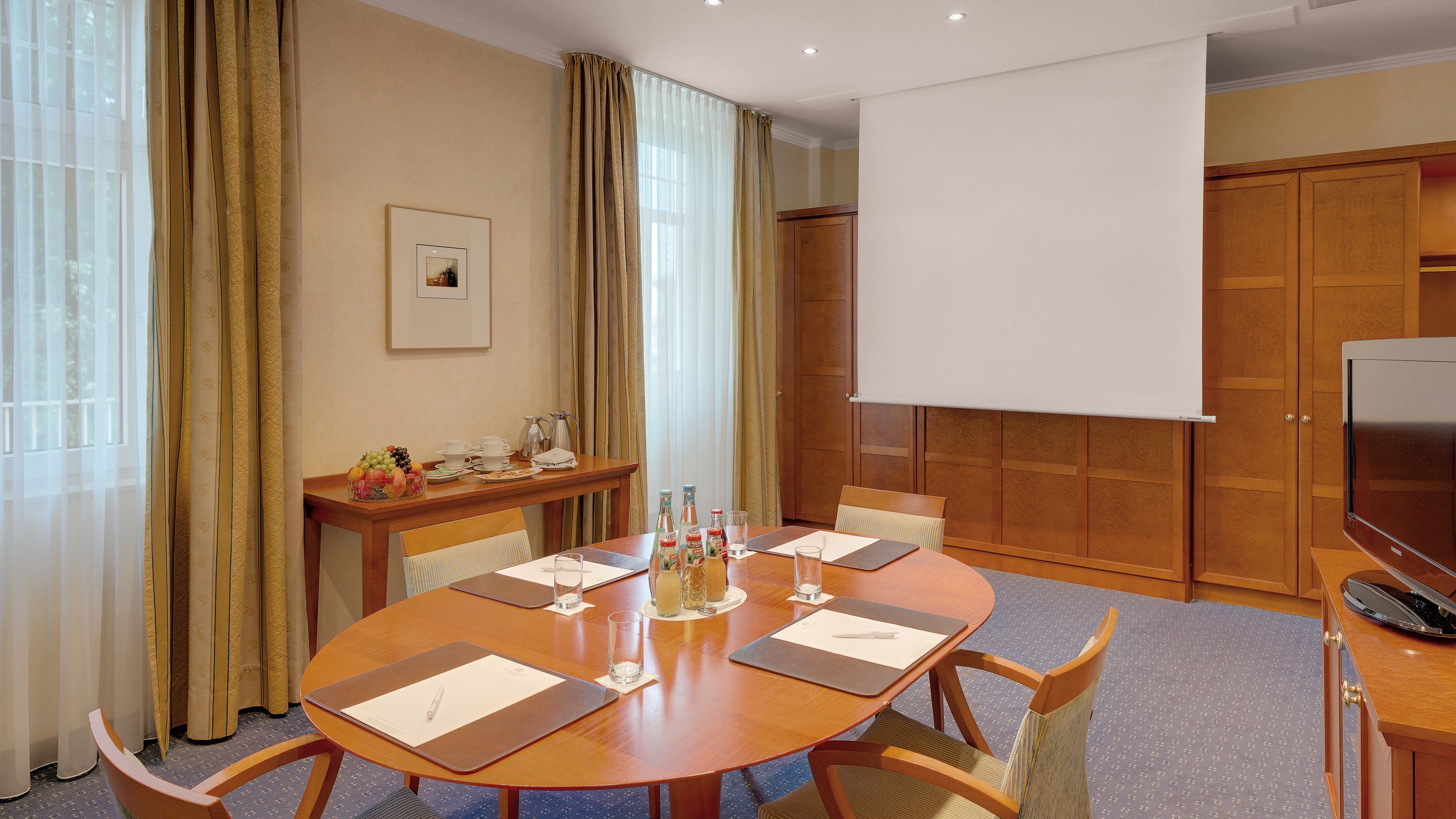 A conference room with a round table, chairs, a projector screen, a television, and a small table with a fruit bowl and coffee pots.