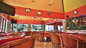 Un restaurante con mesas y sillas de madera, amplias ventanas que dan a palmeras y una decoración en tonos rojo y amarillo.