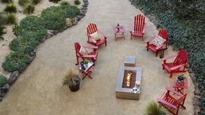 A circular outdoor seating area with red Adirondack chairs and a central fire pit. - Hotel Focus SFO (South San Francisco)
