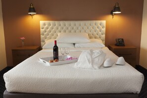 A neatly made bed with a white headboard, a tray with a bottle of wine, glasses, and fruit, and a robe and slippers on the bed. There are two bedside tables with lamps and a phone on one. - Hotel Focus SFO (South San Francisco)