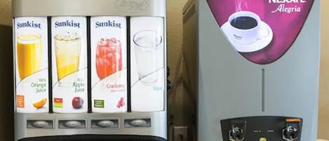 Un dispensador de bebidas con opciones de zumo de naranja, zumo de manzana, zumo de arándano y agua, además de una máquina de café Nescafé.