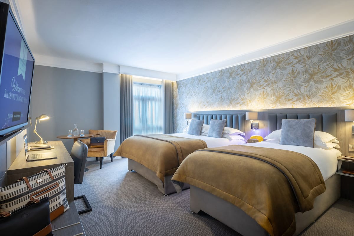 Ein Hotelzimmer mit zwei Betten, einem Schreibtisch mit Lampe, einem Fernseher und einem Fenster mit Vorhängen.
