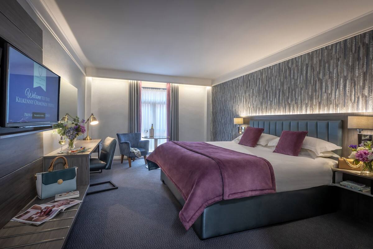 Ein Hotelzimmer mit einem großen Bett, einem Schreibtisch mit Computer, einem an der Wand befestigten Fernseher und einem Fenster mit Vorhängen.