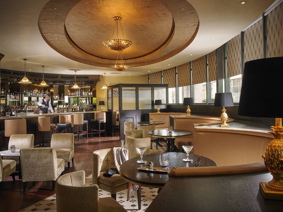 A bar area with a bartender, round tables, and bar stools. There are large windows with patterned curtains and a high ceiling with a circular light fixture.
