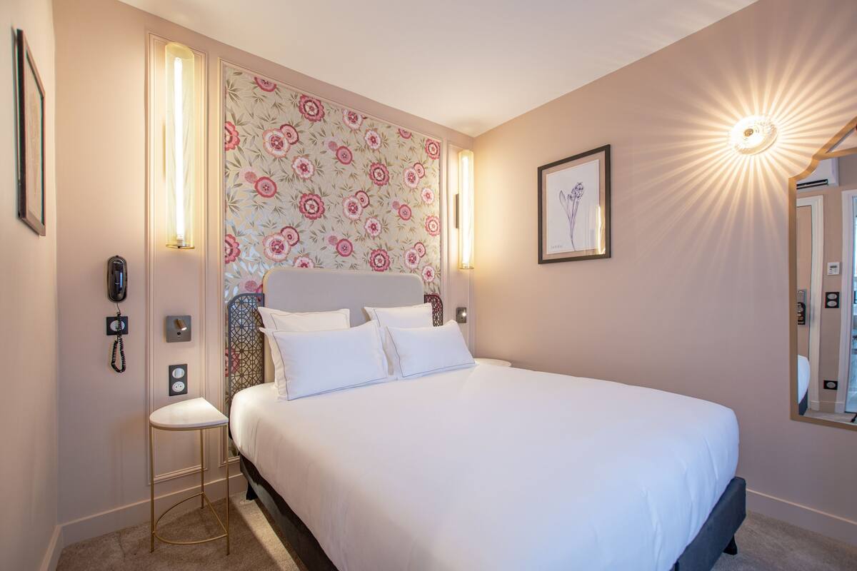 A neatly made bed with white linens, a wall with floral wallpaper, and two wall-mounted light fixtures.