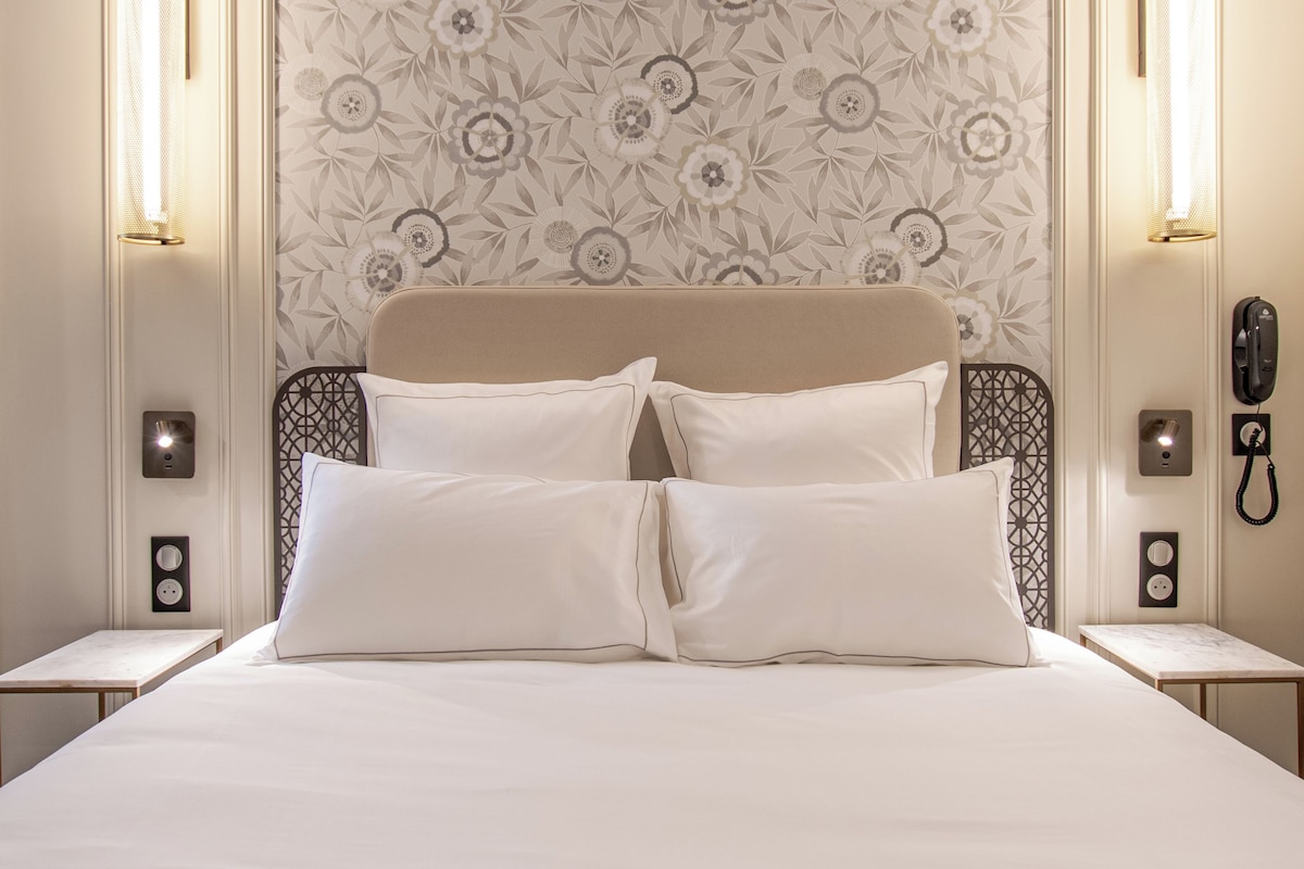 Uma cama bem arrumada com roupa de cama branca, papel de parede estampado com flores e duas luminárias de cabeceira.