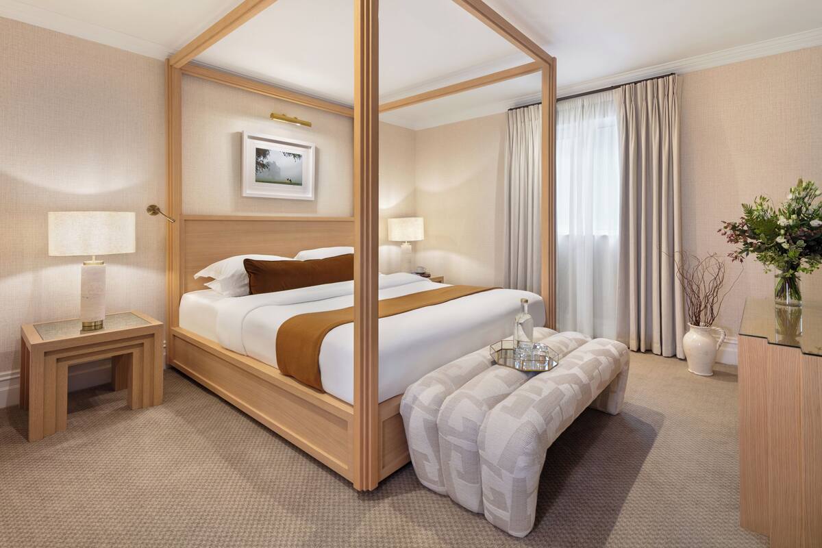A four-poster bed with a wooden frame, two bedside tables with lamps, a bench, a vase with flowers, and a framed picture on the wall.