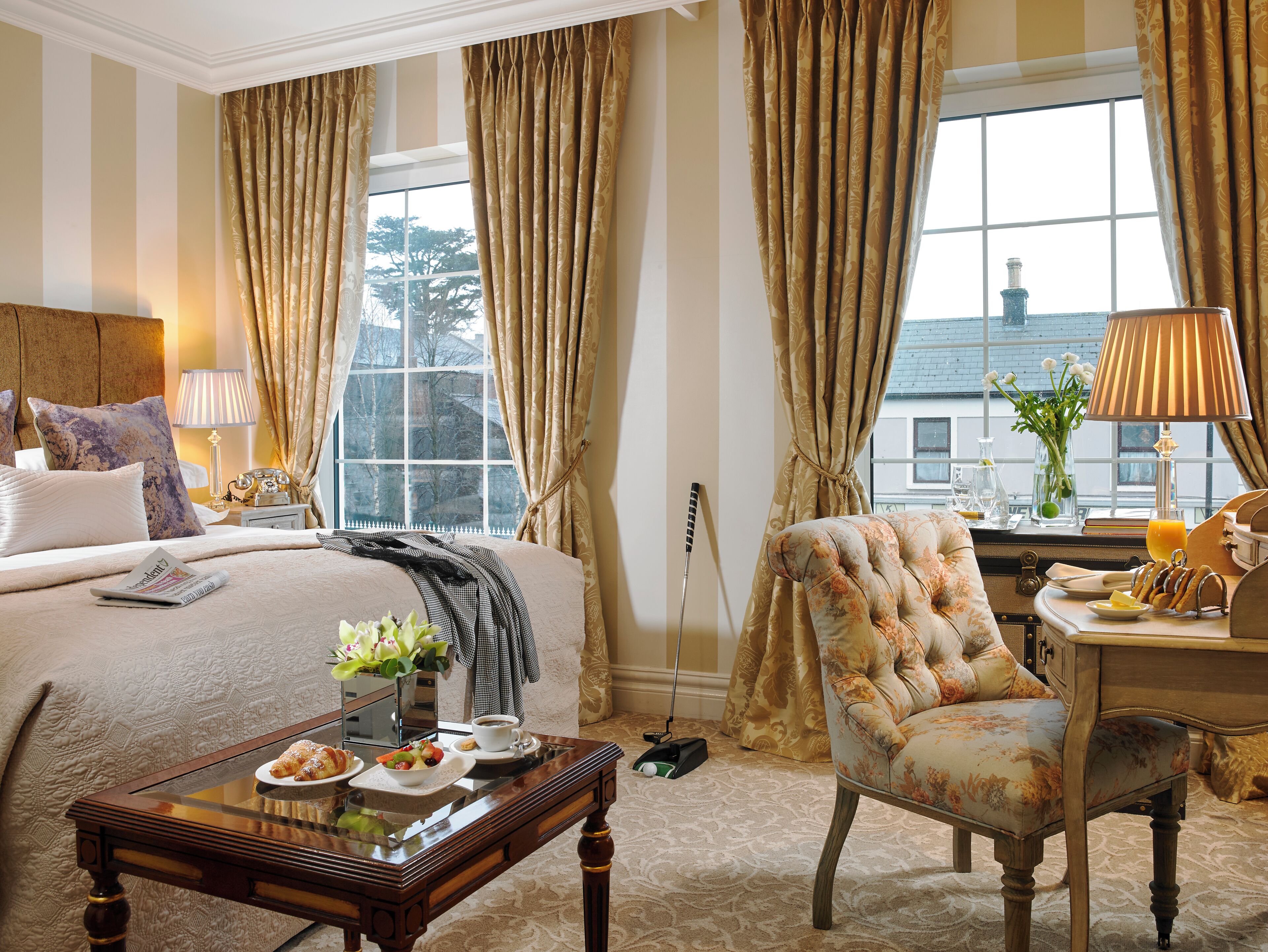 A hotel room with a bed, a chair, a table with breakfast, a lamp, and a window with curtains.