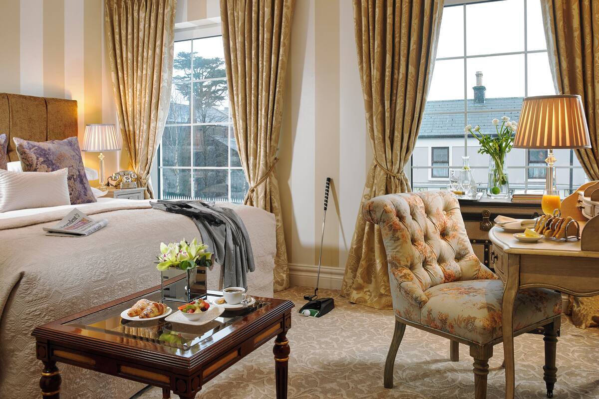 A hotel room with a bed, a chair, a table with breakfast, a lamp, and a window with curtains.