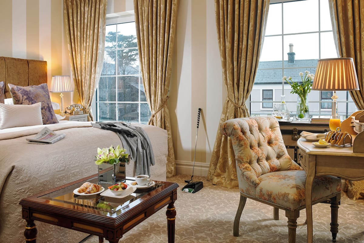 A hotel room with a bed, a chair, a table with breakfast, a lamp, and a window with curtains.