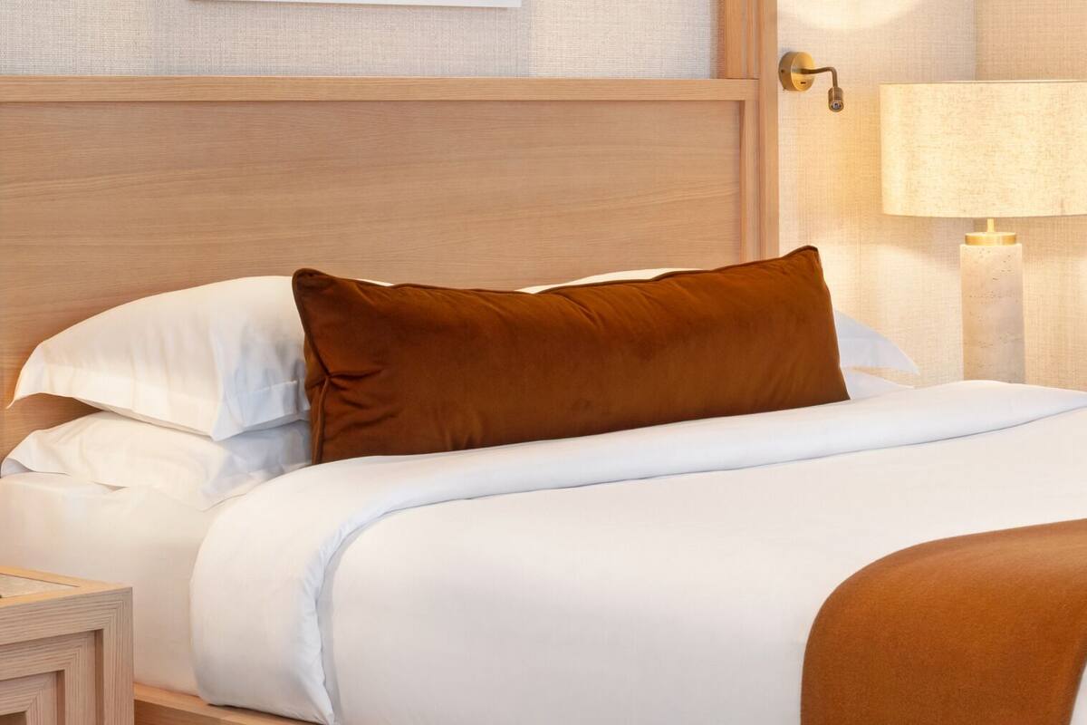 A neatly made bed with white bedding and a brown accent pillow, a wooden headboard, a bedside lamp, and a nightstand.