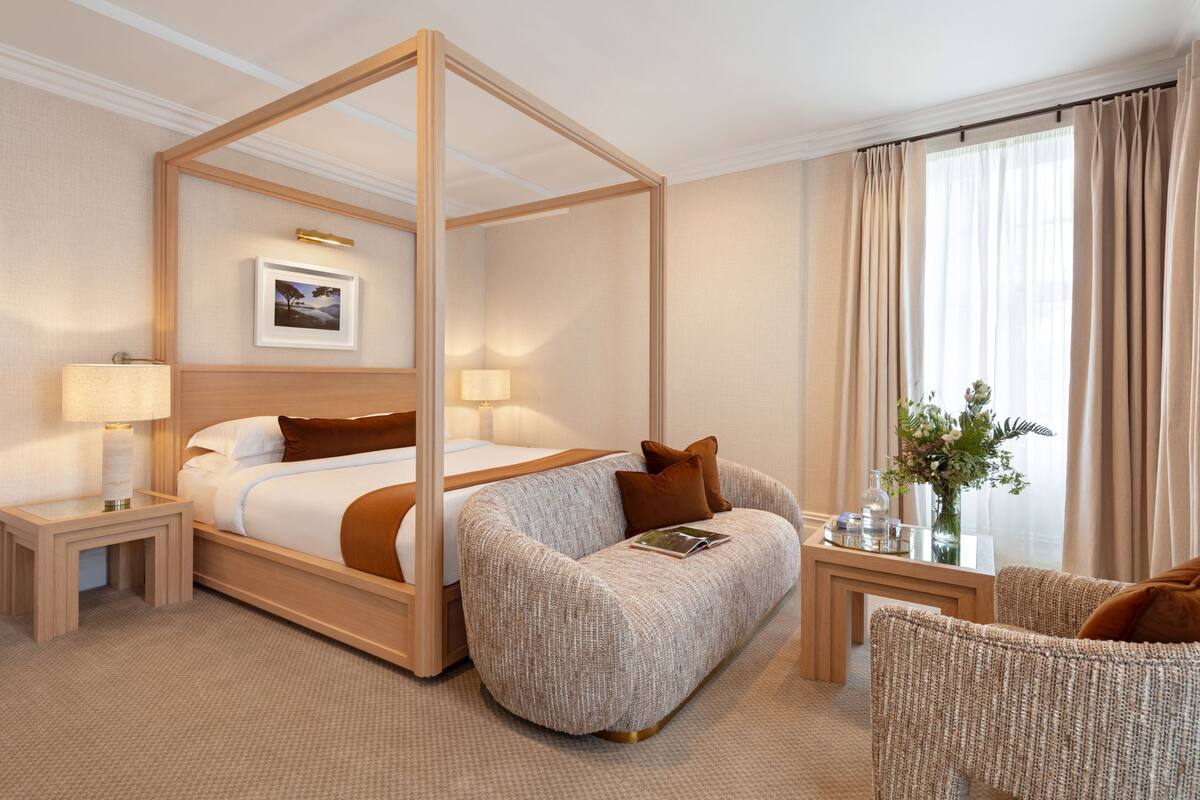 A four-poster bed with a wooden frame, a bedside table with a lamp, a sofa, and a small table with a vase of flowers.