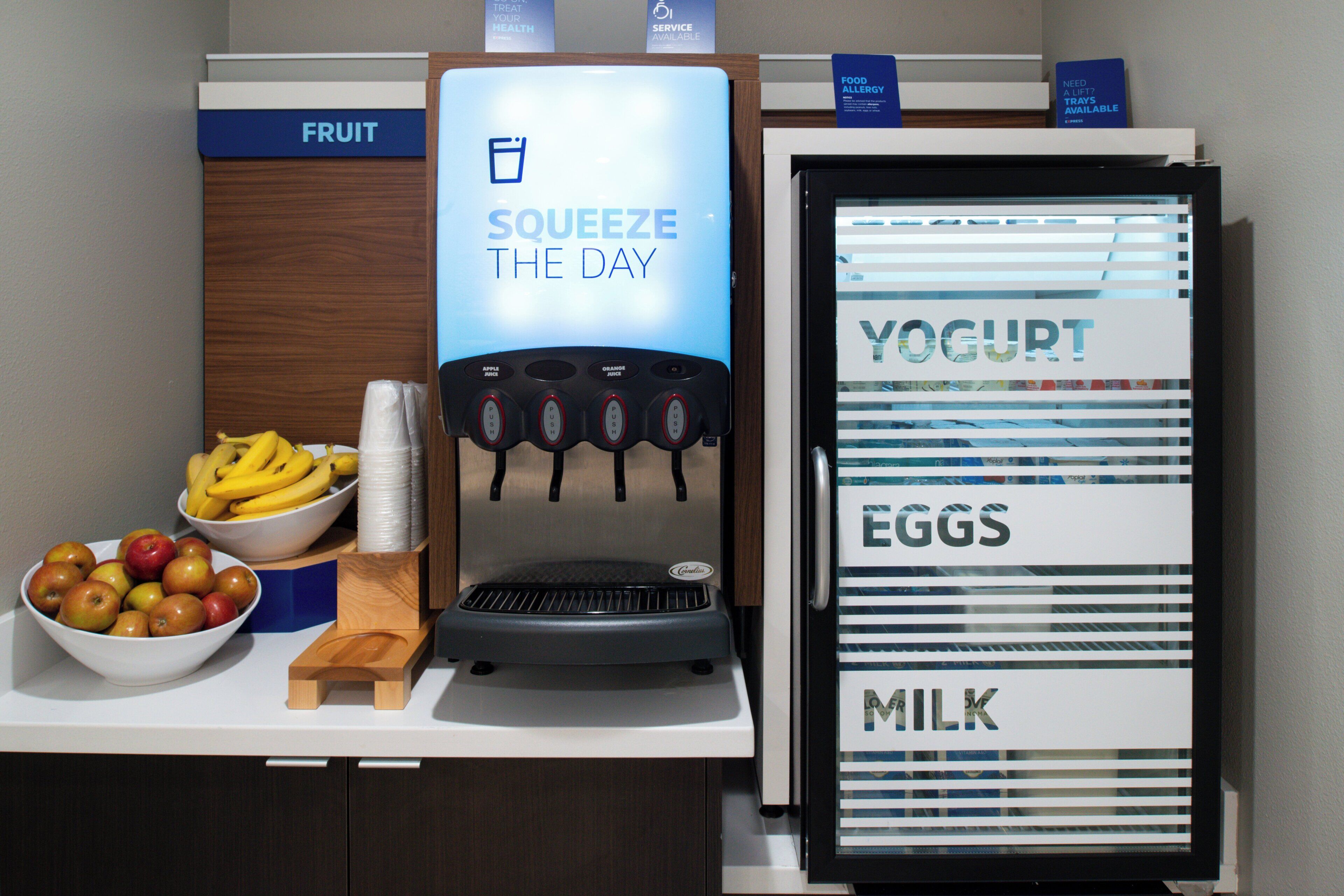 A self-serve station with a beverage dispenser, a bowl of fruit, and a refrigerated display for yogurt, eggs, and milk.