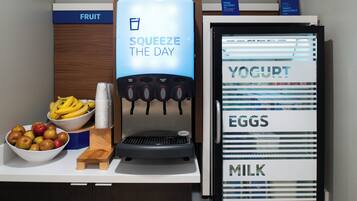 A self-serve station with a beverage dispenser, a bowl of fruit, and a refrigerated display for yogurt, eggs, and milk.