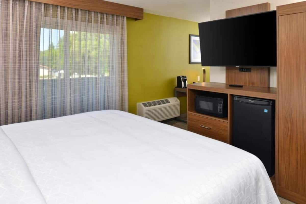 A hotel room with a bed, a television, a microwave, a refrigerator, and an air conditioning unit.