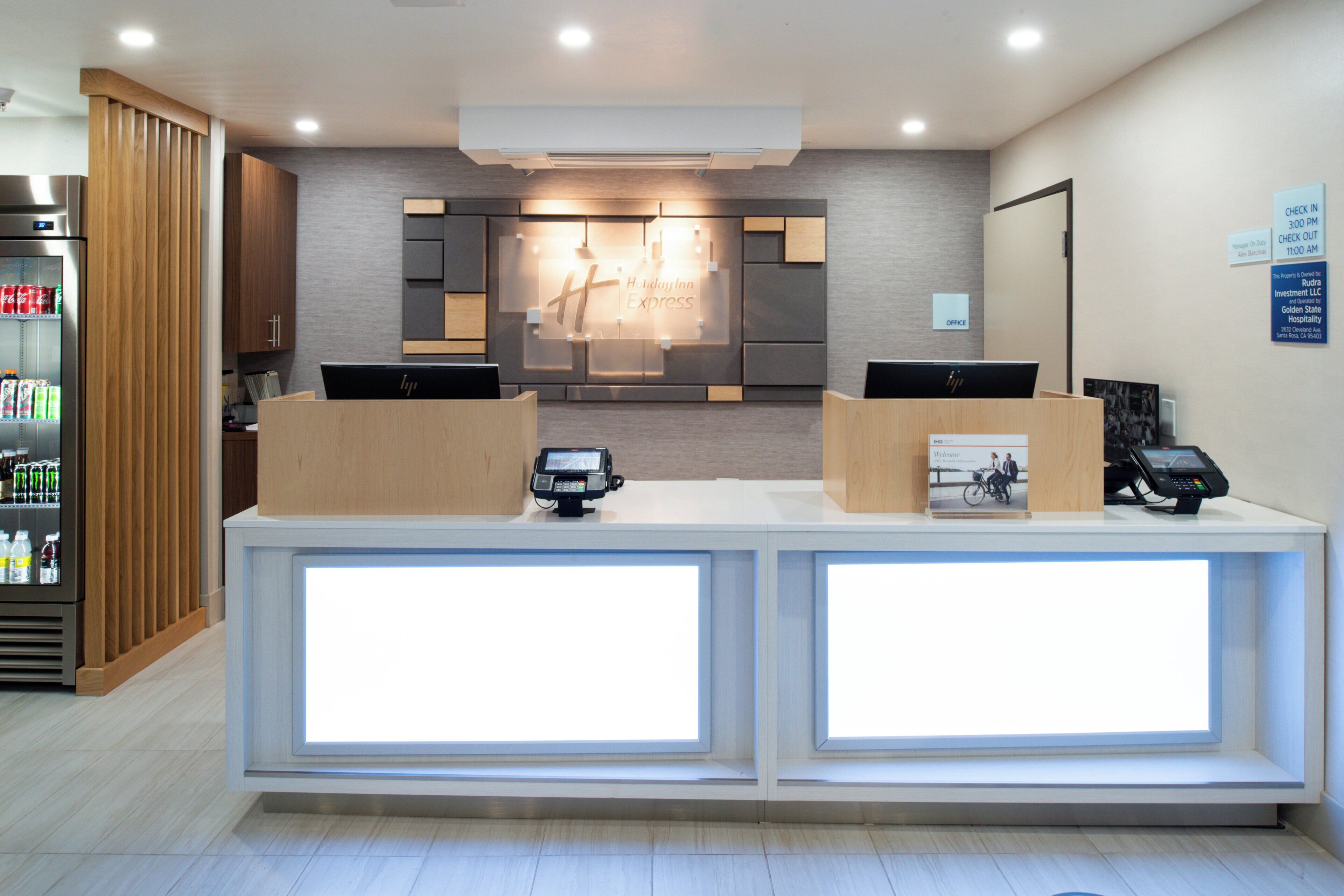 A reception desk with two computer monitors, a refrigerator with beverages, and a wall-mounted sign indicating check-in and check-out times.