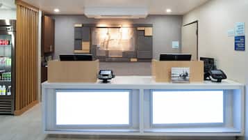 A reception desk with two computer monitors, a refrigerator with beverages, and a wall-mounted sign indicating check-in and check-out times.