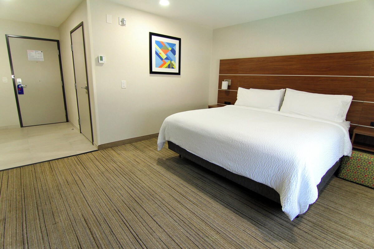 A hotel room with a large bed, a wooden headboard, a framed picture on the wall, and a door to the left.
