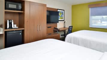 Habitación de hotel con dos camas, kitchenette con microondas, cafetera y refrigerador pequeño, escritorio con computadora y teléfono, y televisor sobre el escritorio.