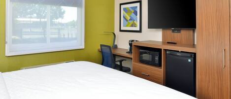 A hotel room with a bed, a desk with a computer, a television, and a refrigerator.