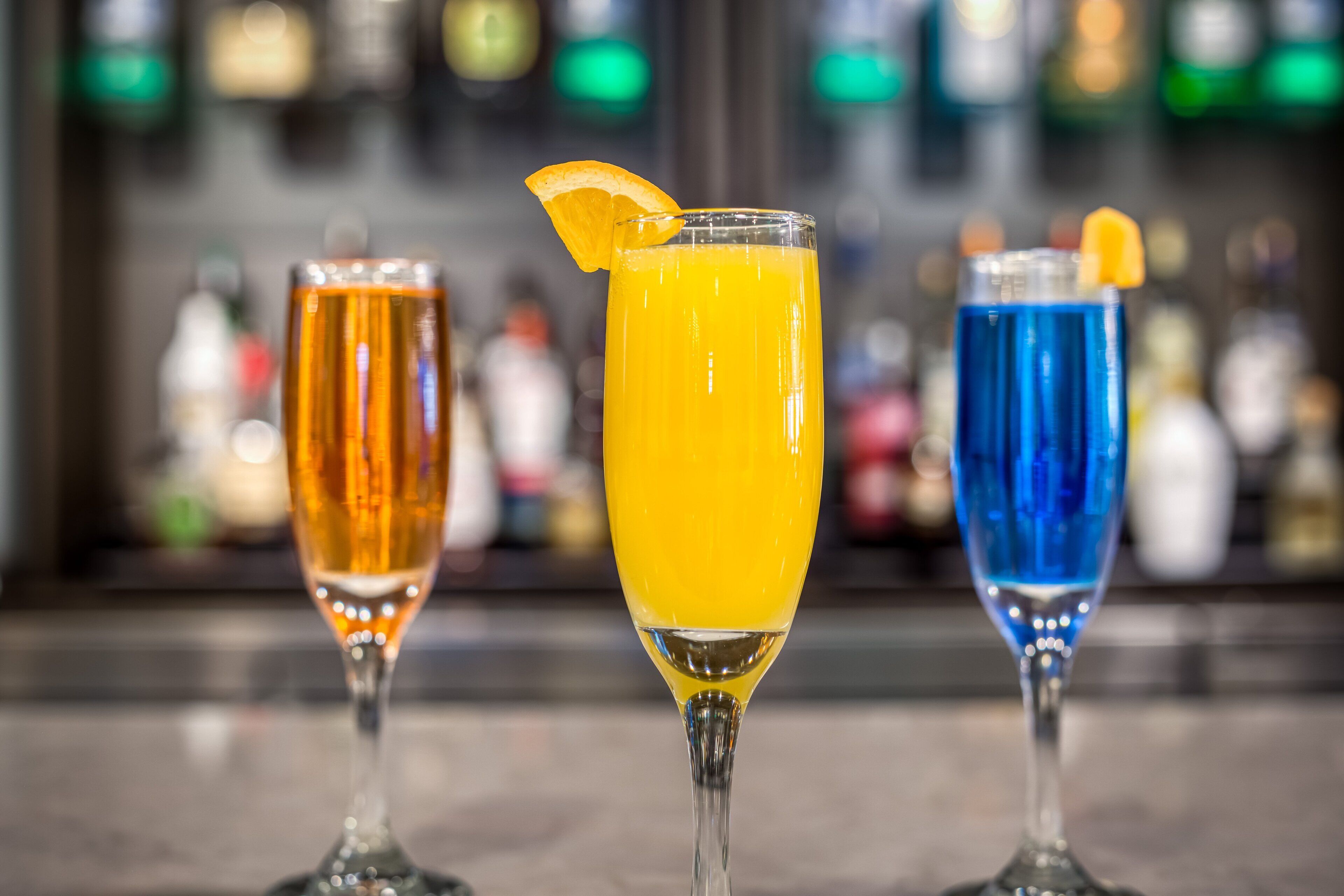 Trois verres de boissons sur le comptoir d’un bar, avec un arrière-plan flou composé de bouteilles.