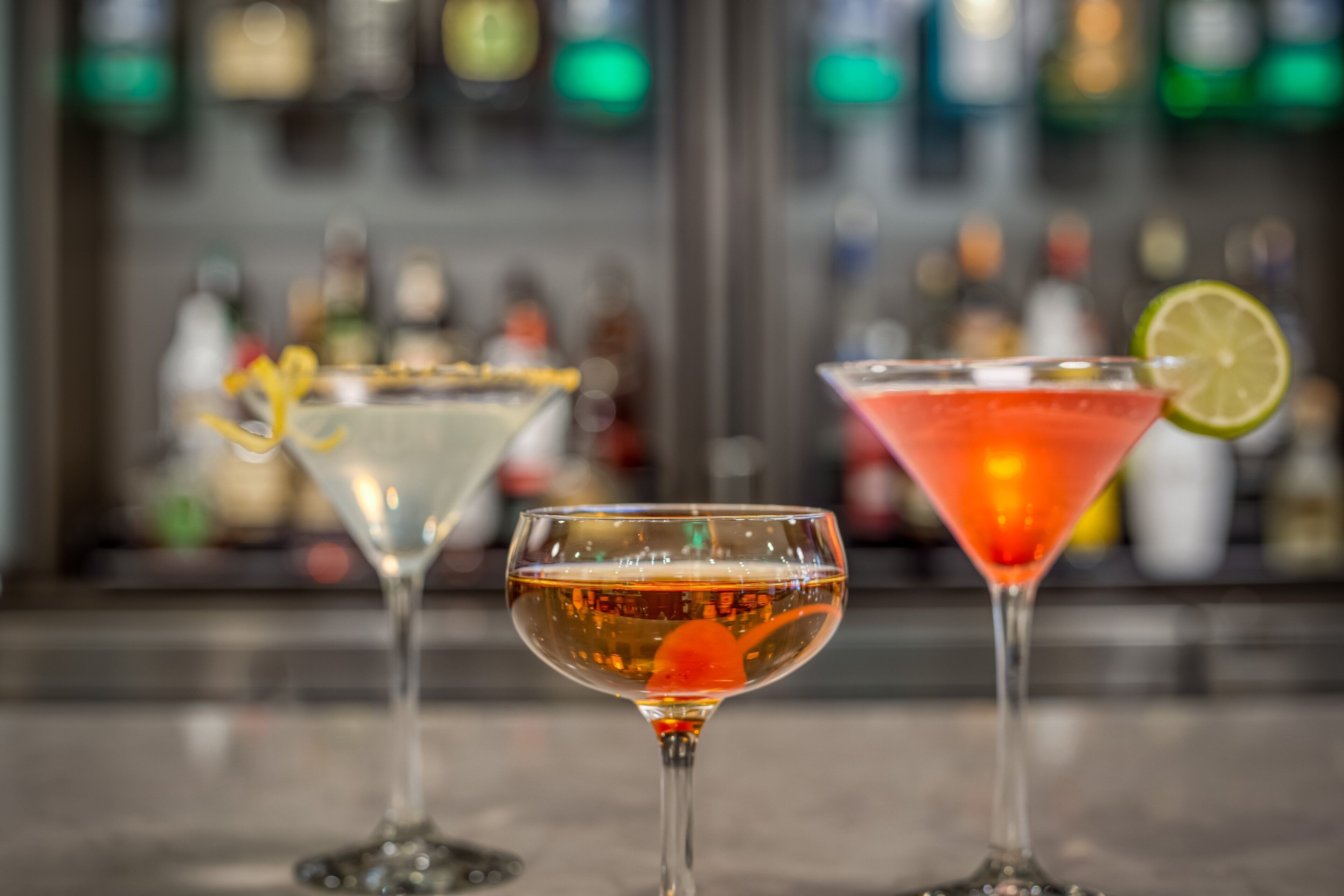 Three cocktails on a bar counter with a blurred background of bottles.