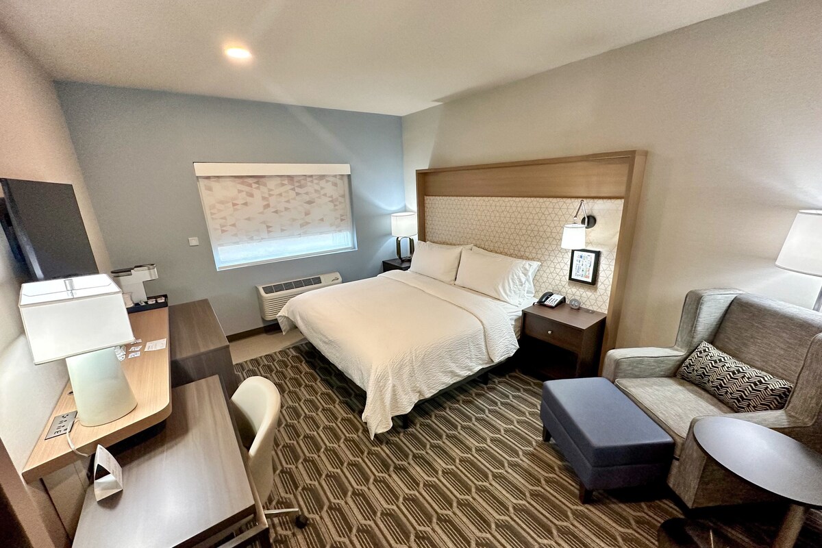 A hotel room with a large bed, a desk with a lamp, a chair, a sofa, and a bedside table with a phone and a lamp.