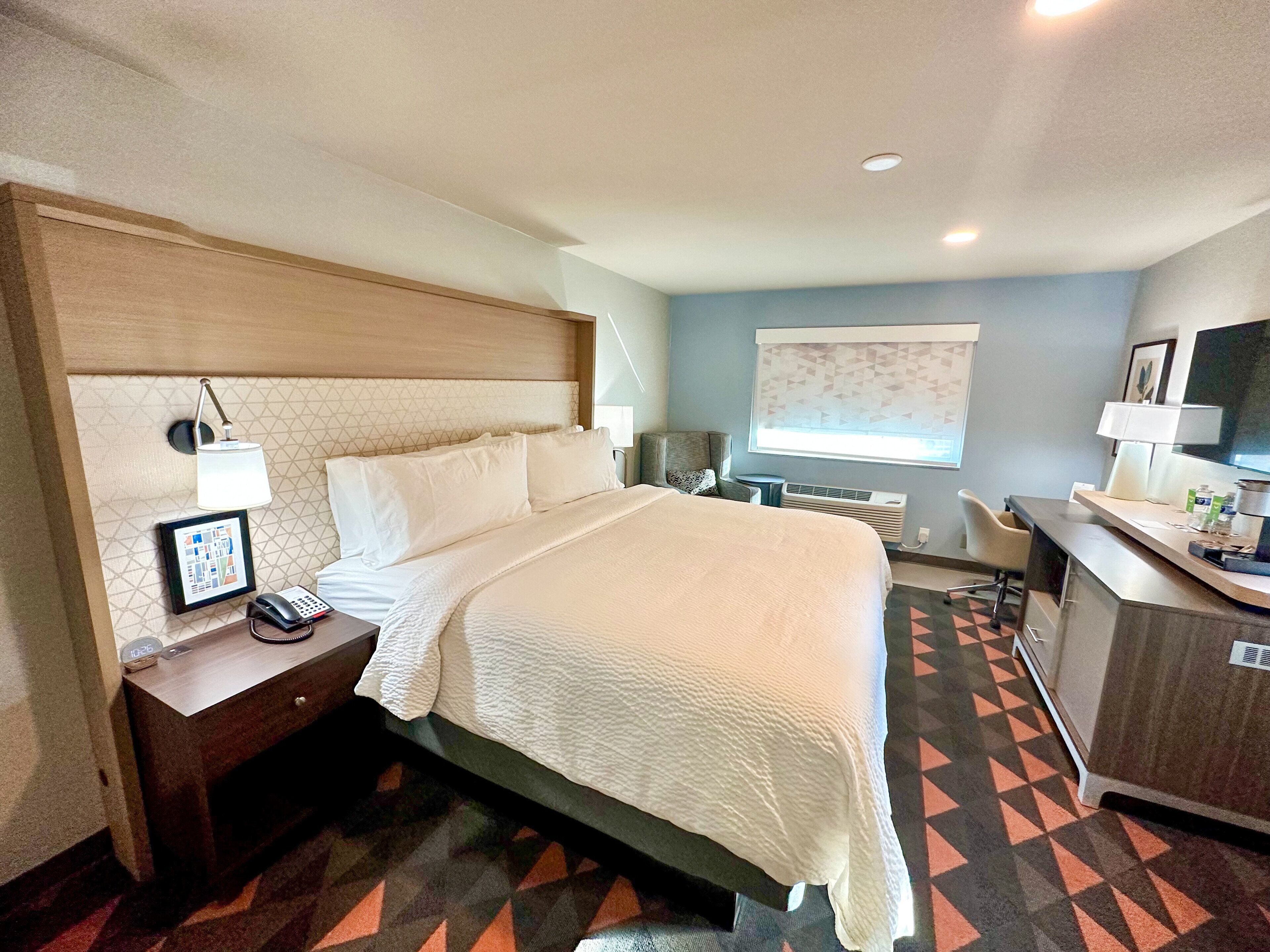 A hotel room with a bed, bedside table, lamp, and a desk with a chair.
