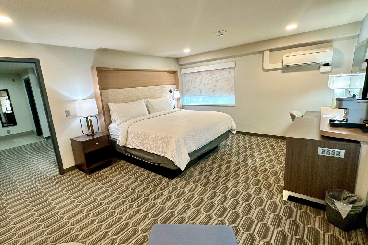 A hotel room with a bed, a desk, a trash bin, and a window with blinds.