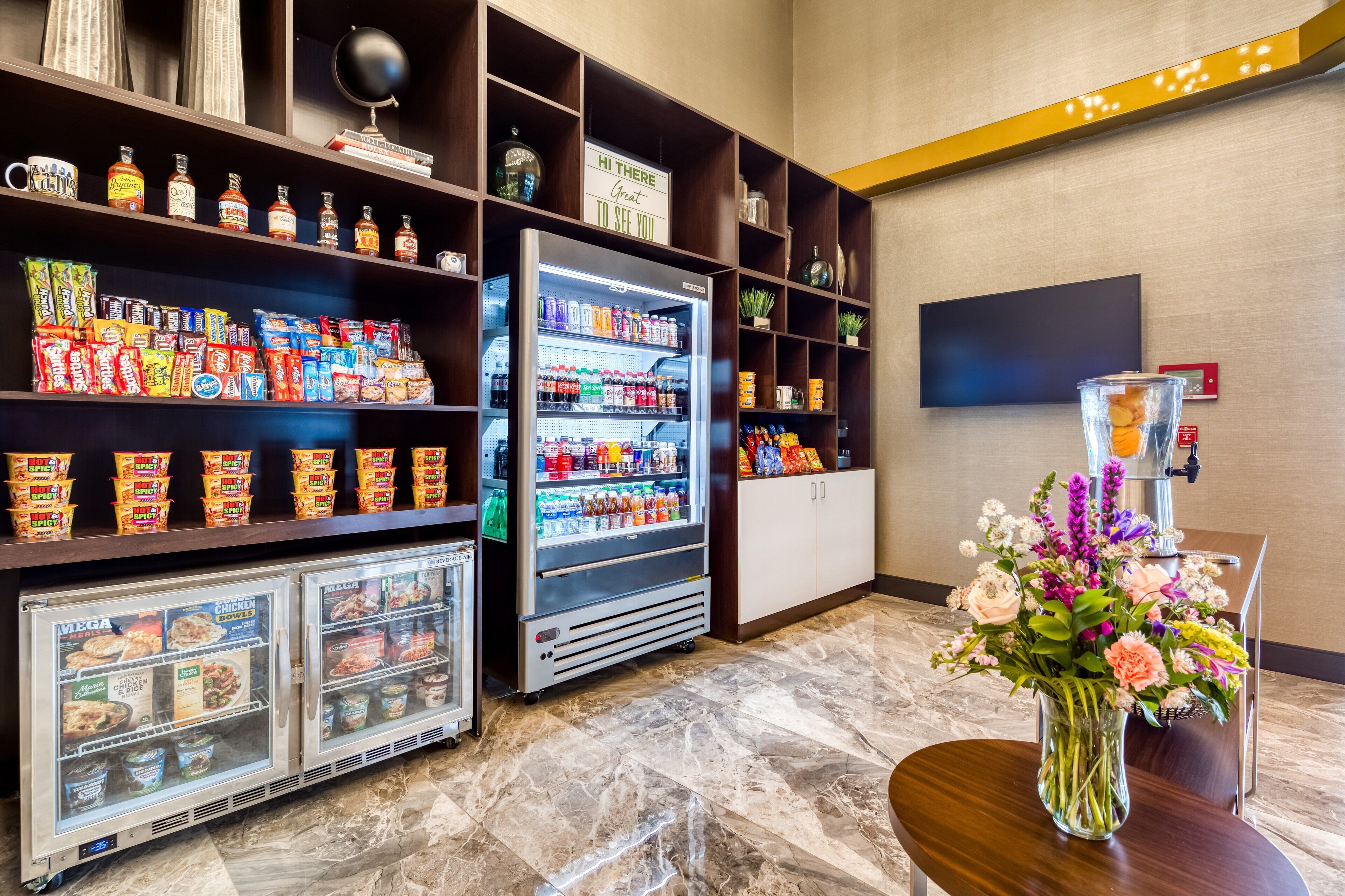 A hotel lobby with a self-service beverage fridge, snack dispensers, and a floral arrangement on a table.