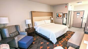 A hotel room with a large bed, a desk, a chair, a small table, and a blue ottoman.