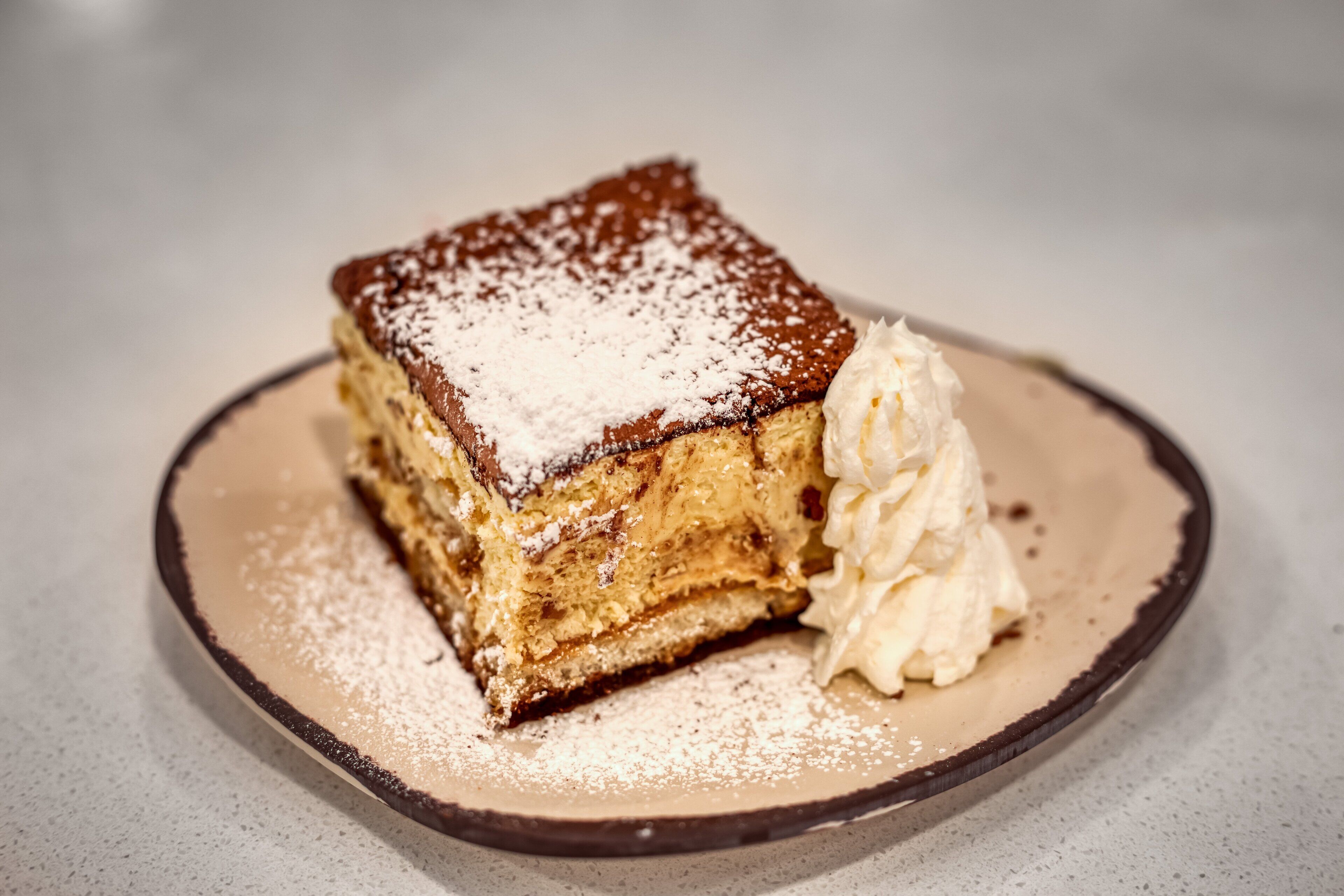 Una porció de postre en capes sobre un plat, amb sucre glas i una culleradeta de crema batuda.