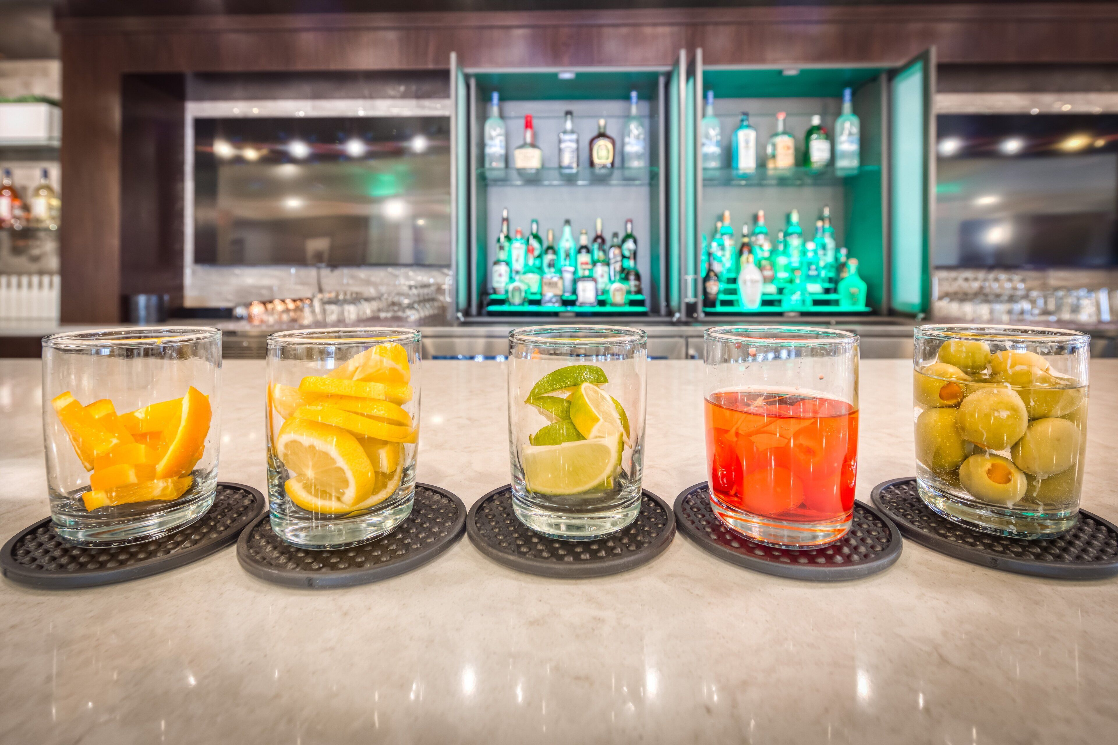 A bar area with five glasses of different beverages, each with a slice of fruit, and a background of shelves with various bottles.