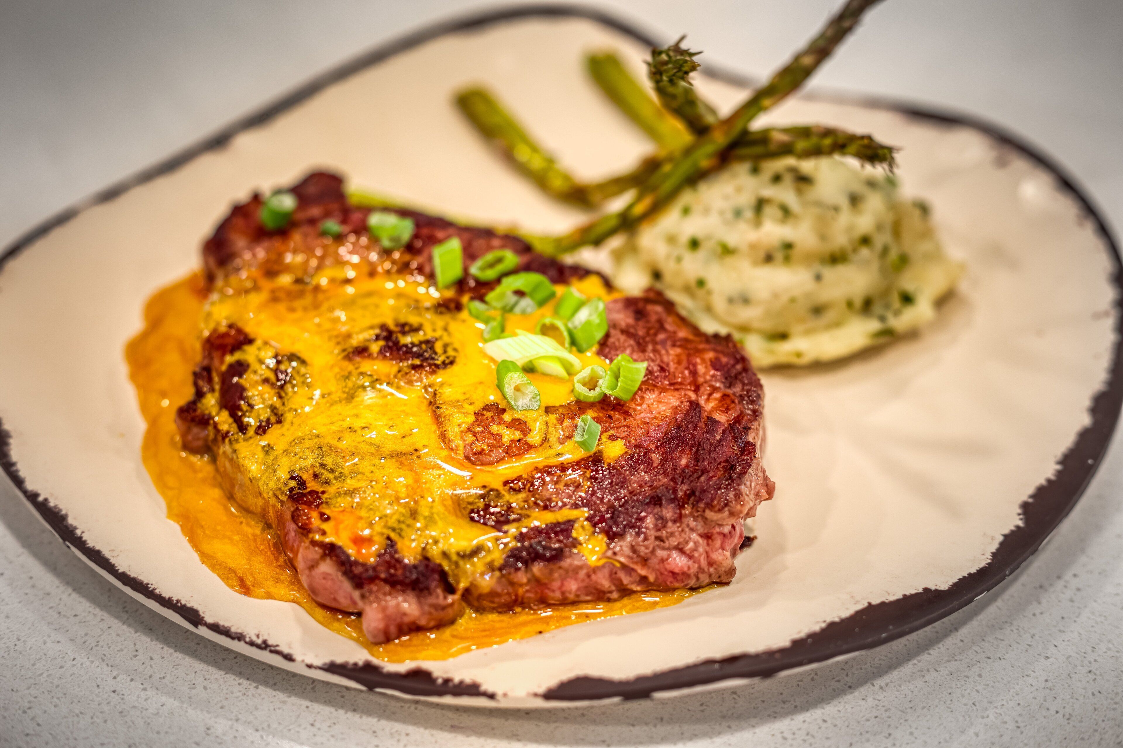 A steak with melted cheese and green onions on a plate with asparagus and mashed potatoes.
