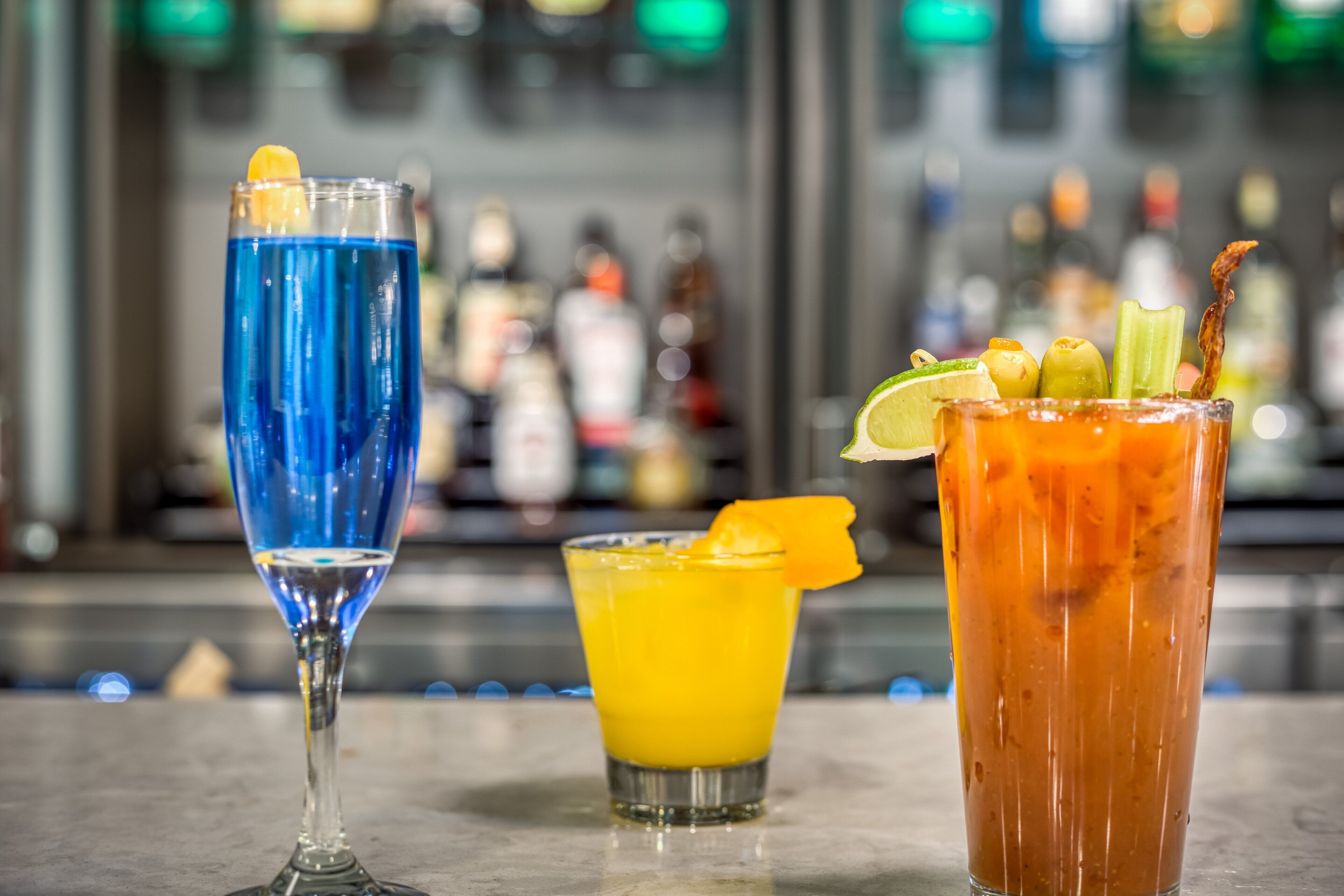 Three cocktails on a bar counter with a blurred background of bottles.