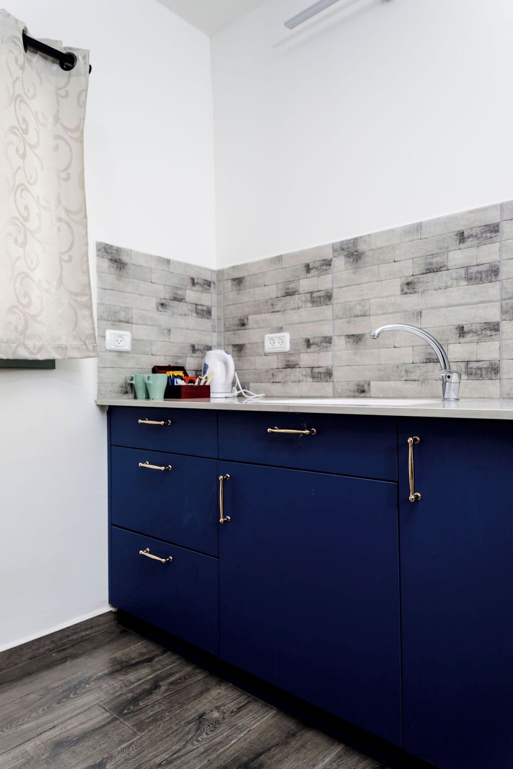 Uma cozinha com design moderno, apresentando um painel de azulejos cinza, uma pia de aço inoxidável e armários azul escuro com puxadores dourados.