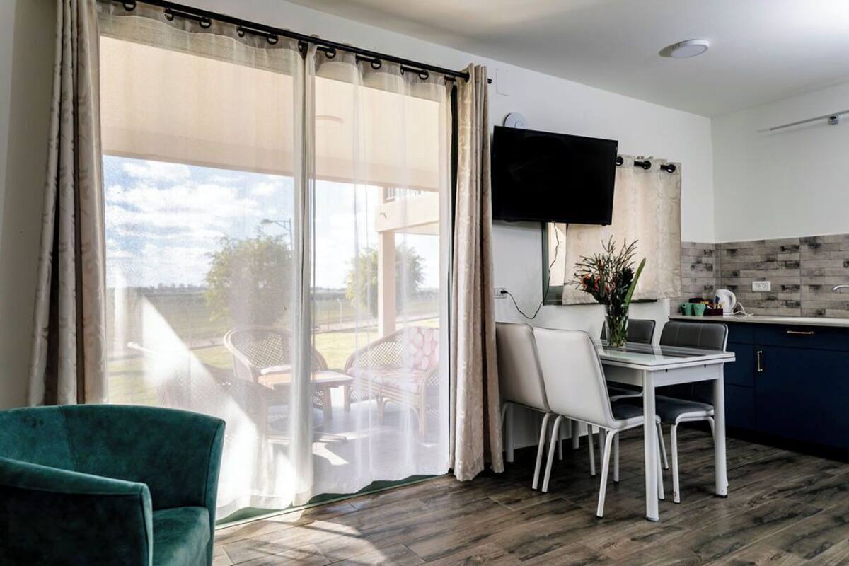 A modern kitchen with a flat-screen TV, a dining area with a table and chairs, and a view through large windows.