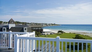 A coastal view from a balcony with a white railing, overlooking a beach and ocean.