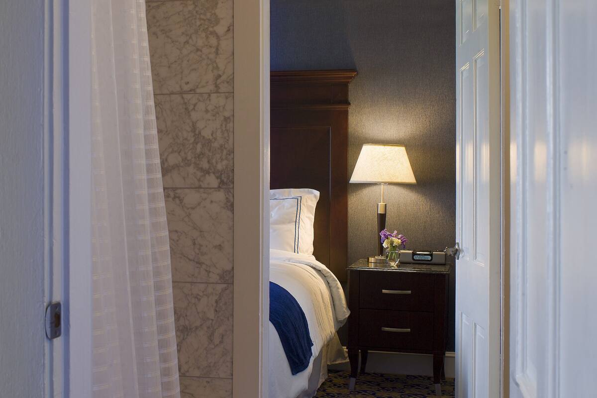 Un couloir étroit mène à une chambre à coucher qui contient un lit, une table de chevet et une lampe.