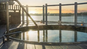 Un jacuzzi con plataforma de madera y baranda metálica, con vista a un cuerpo de agua al atardecer.